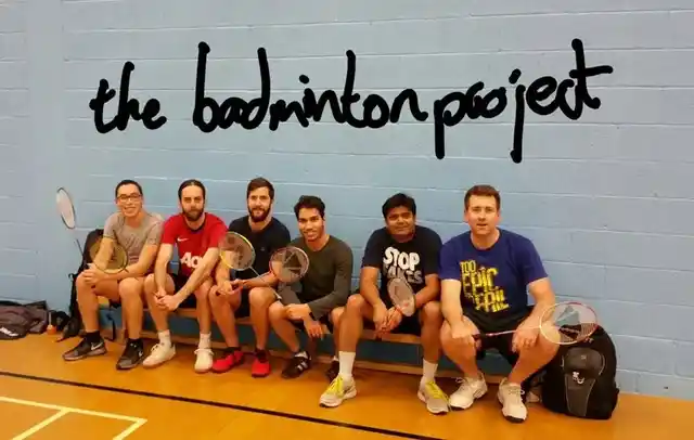 Six badminton players sitting together with rackets beneath The Badminton Project wall sign.