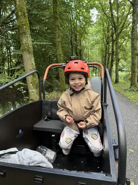 A child wearing a red helmet sitting in a bicycle trailer on a leafy path.