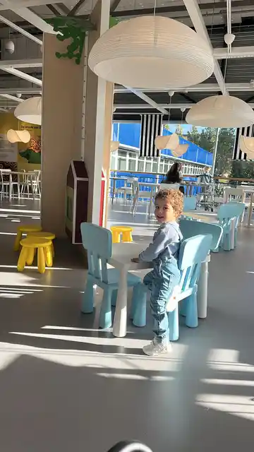 A smiling child standing beside a small table and pale blue chairs in a sunny café.