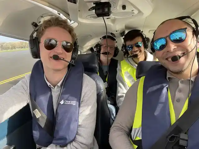 Four people wearing aviation headsets and life jackets inside a light aircraft.