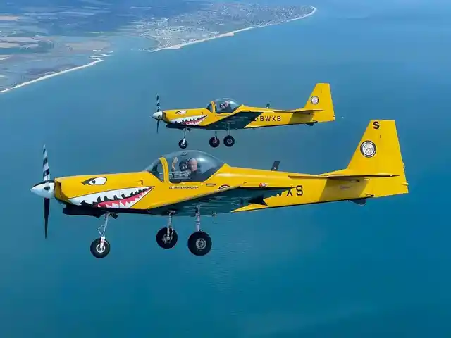 Two yellow light aircraft flying alongside each other above the sea and coastline.