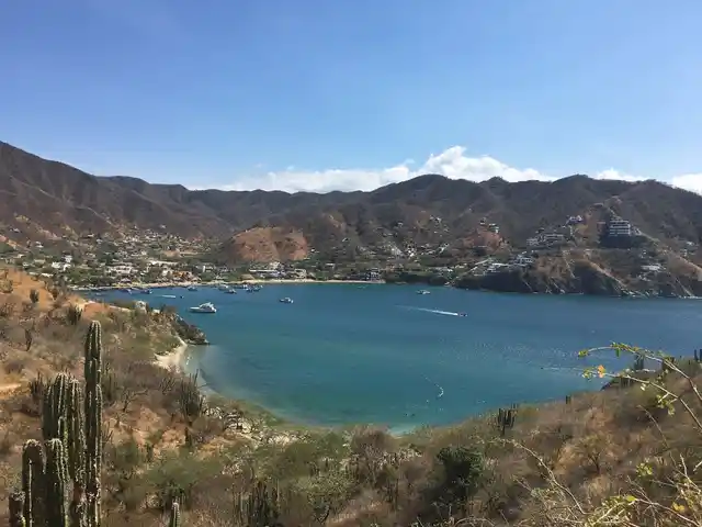A blue bay with boats, surrounded by dry hills, cacti and hillside buildings.
