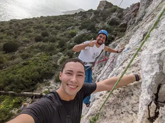 Two climbers secured with ropes on a rocky cliff overlooking trees and the sea.